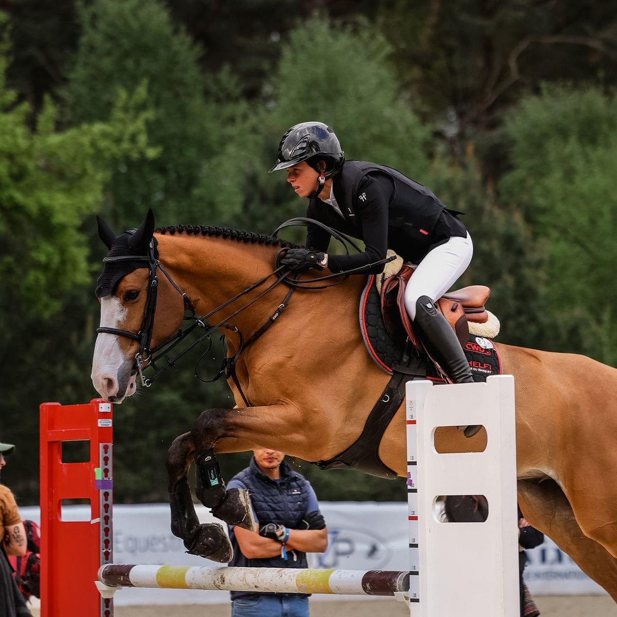 Equestrian rider on a brown horse jumping over a white obstacle during a competition.