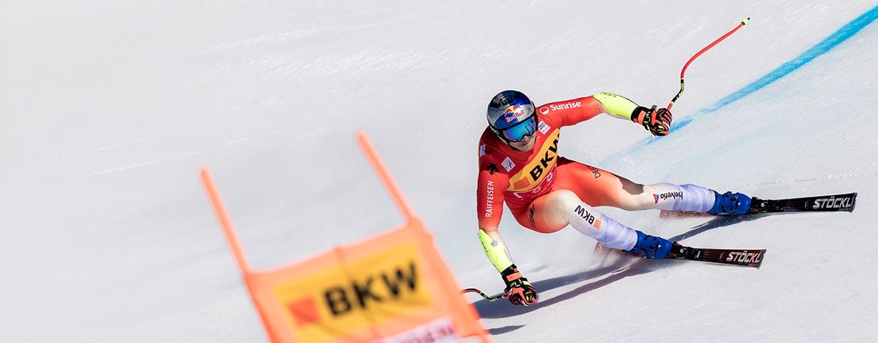 A skier in a red suit carves a turn past an orange gate on a snowy slope.