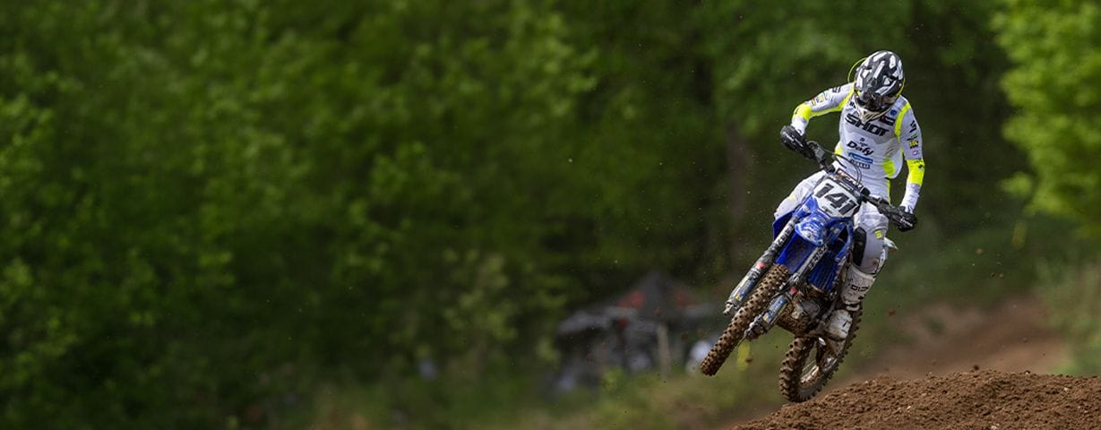 Motocross rider jumps a blue dirt bike on a track with green trees.