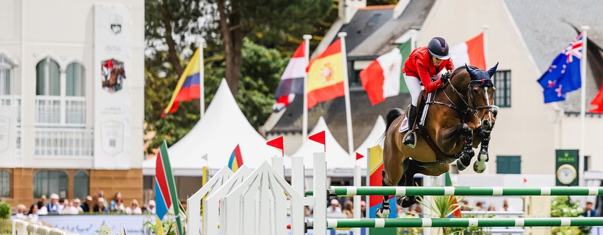 Equestrian rider in red on a dark horse jumping over a white obstacle at a competition.