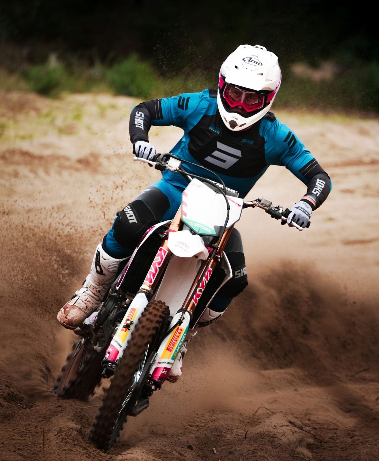 A motocross rider in protective gear speeds through a dirt track, kicking up dust.