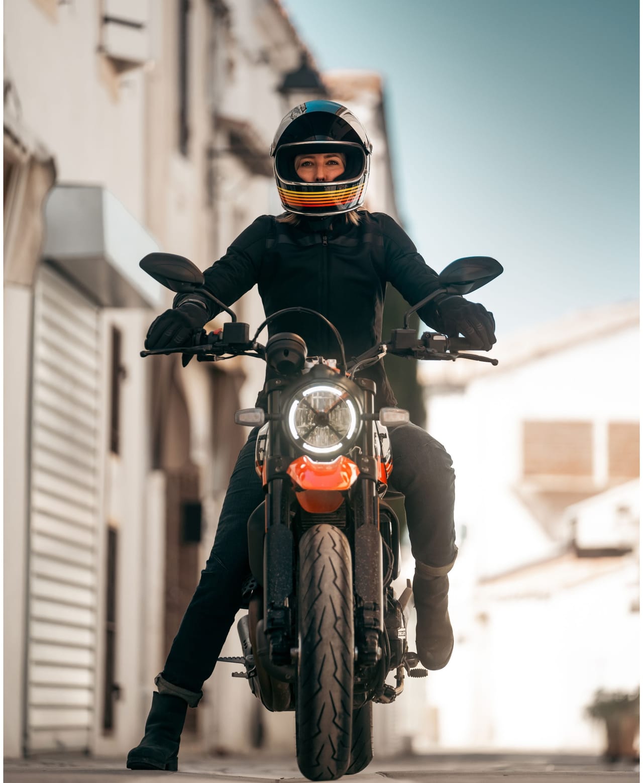 Person in black helmet and gear sits on an orange motorcycle, looking directly at viewer.