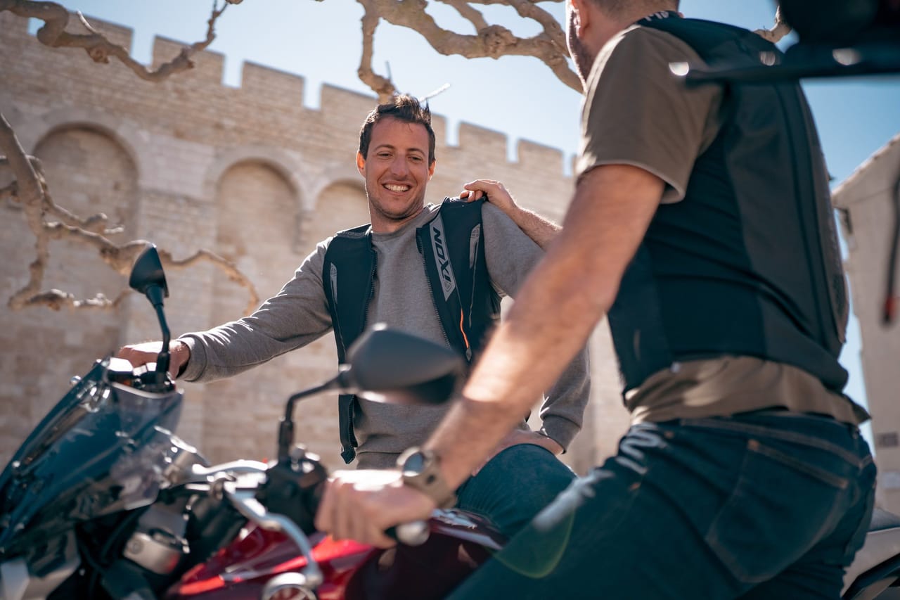 Two smiling men chatting next to their motorcycles in front of an ancient stone wall.