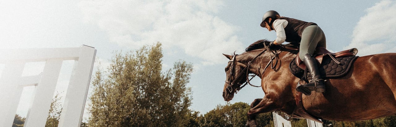 A rider on a brown horse jumping over a white obstacle outdoors.