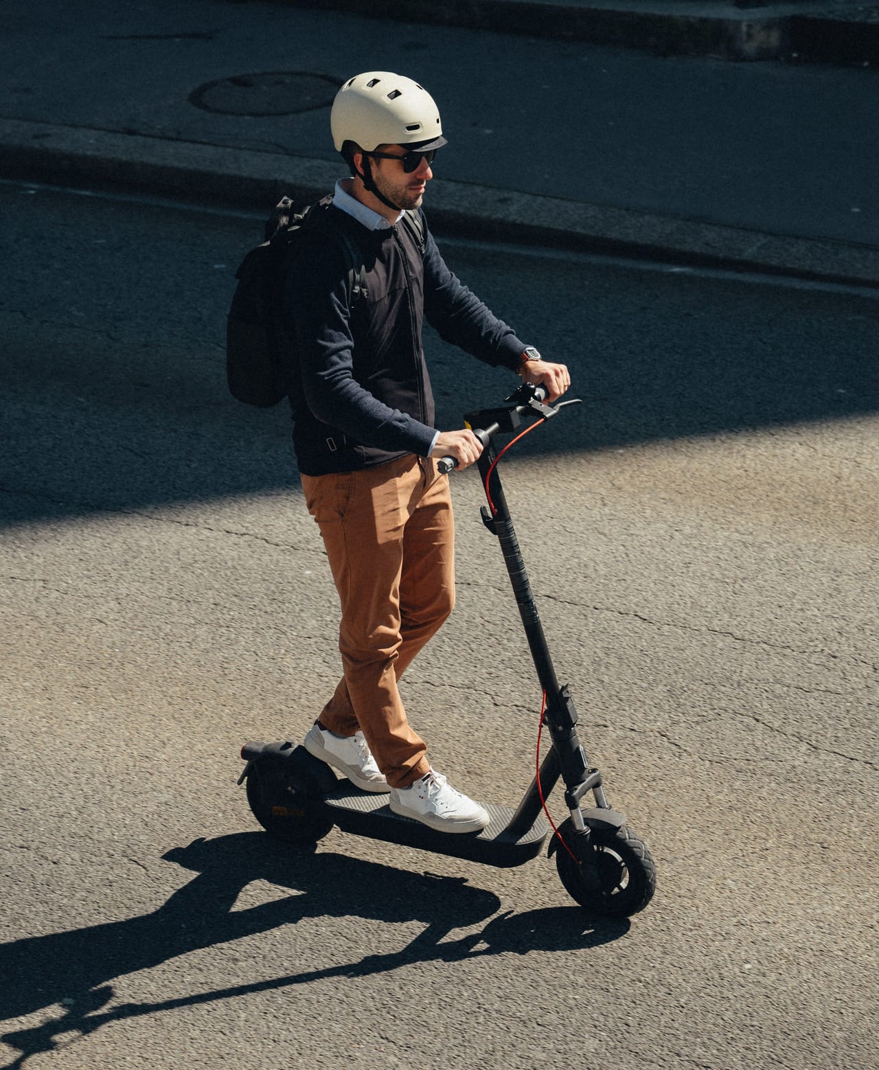 A man wearing a helmet and backpack rides an electric scooter on a paved street.