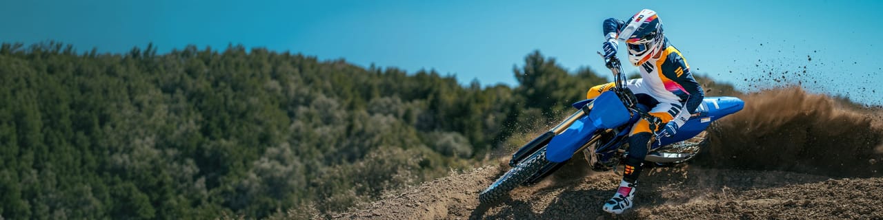 Rider on a blue and yellow dirt bike speeds along a dirt track by green hills.