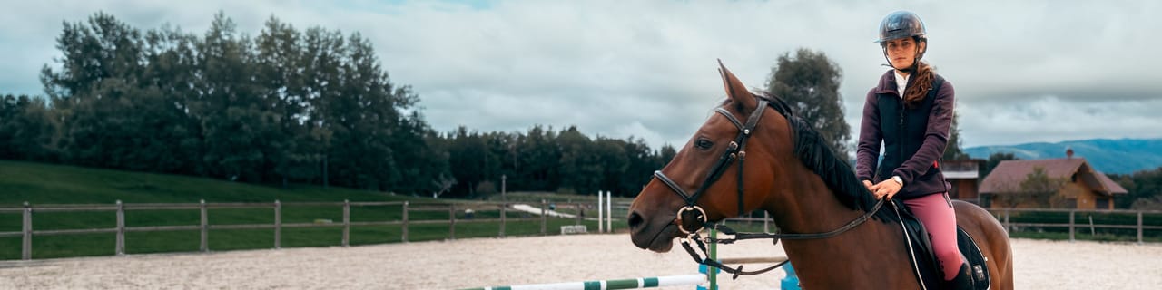 A young rider wearing a helmet sits on a brown horse in an outdoor riding arena.