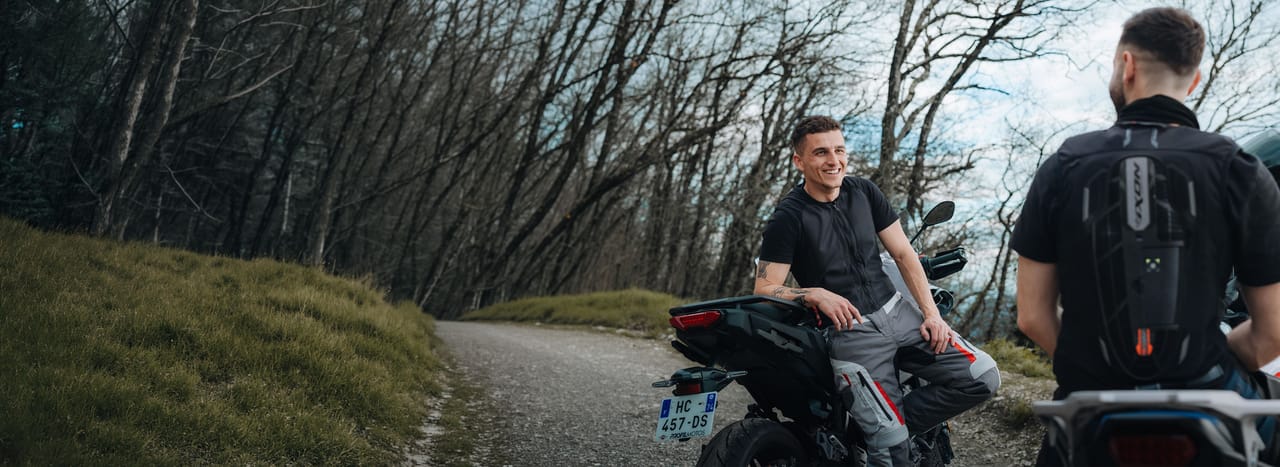Two men on motorcycles on a winding road through a sparse, wooded area.