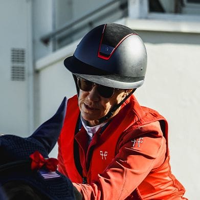 Person wearing a black equestrian helmet, sunglasses, and a bright orange jacket.