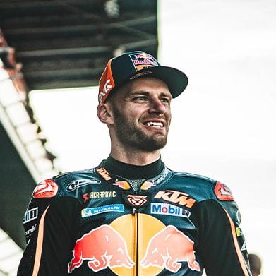 Male motorcycle racer smiling in a racing suit and an American flag cap.