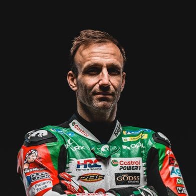 A male motorcycle racer in a green and white suit looks forward against a dark background.